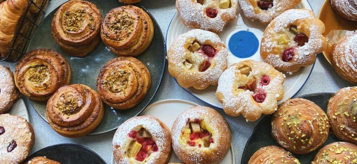 Widok z góry na różnorodne rzemieślnicze wypieki: drożdżówki z owocami i kruszonką, cynamonki oraz bułeczki z pistacjami ułożone na ceramicznych talerzach.